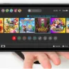 Una mano tiene una Nintendo Switch 2 con lo schermo che mostra la schermata di selezione dei profili utente, con diverse icone colorate e nickname, a simboleggiare la personalizzazione per ogni giocatore.